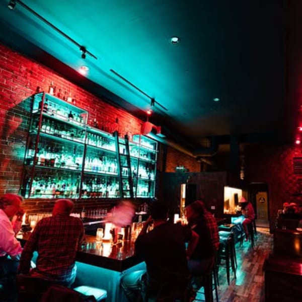 Patrons sit at a bar with a brick wall and shelves of bottles illuminated by blue and red lights.