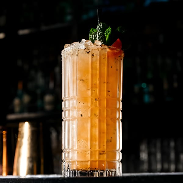 A tall, orange cocktail with crushed ice, mint, and a fruit garnish, against a dark bar background.