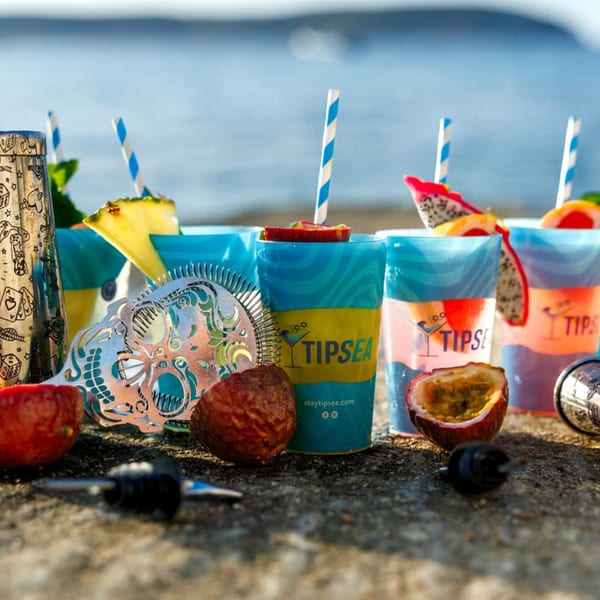 Tipsea branded cups with tropical drinks, fruit garnishes, and bar tools on a stone surface by the sea.