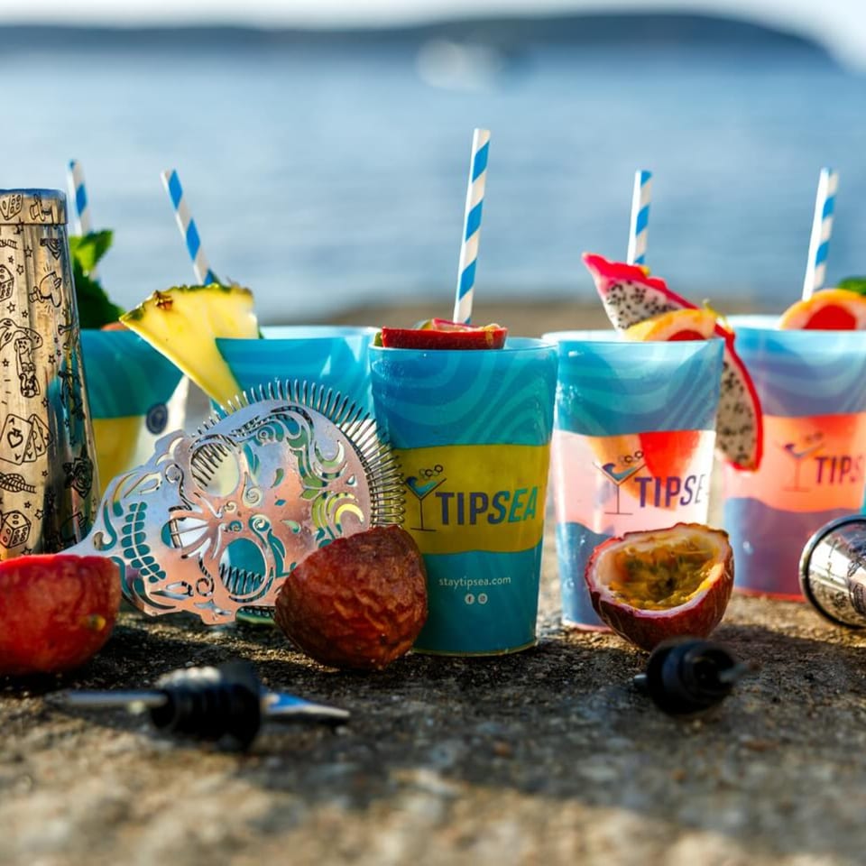 Tipsea branded cups with tropical drinks, fruit garnishes, and bar tools on a stone surface by the sea.