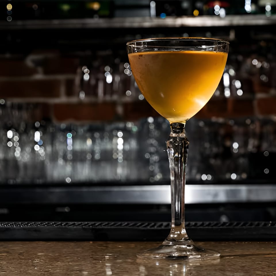 A golden cocktail in a stemmed glass sits on a bar with a brick wall and blurred background.