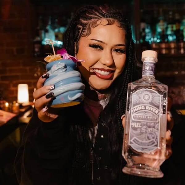 A smiling woman holding a blue tiki cocktail and a bottle of Ming River baijiu.