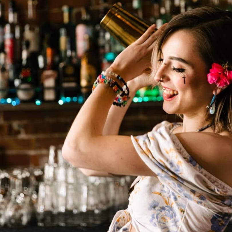 A smiling bartender with a flower in her hair shakes a cocktail shaker at Tipsea bar.